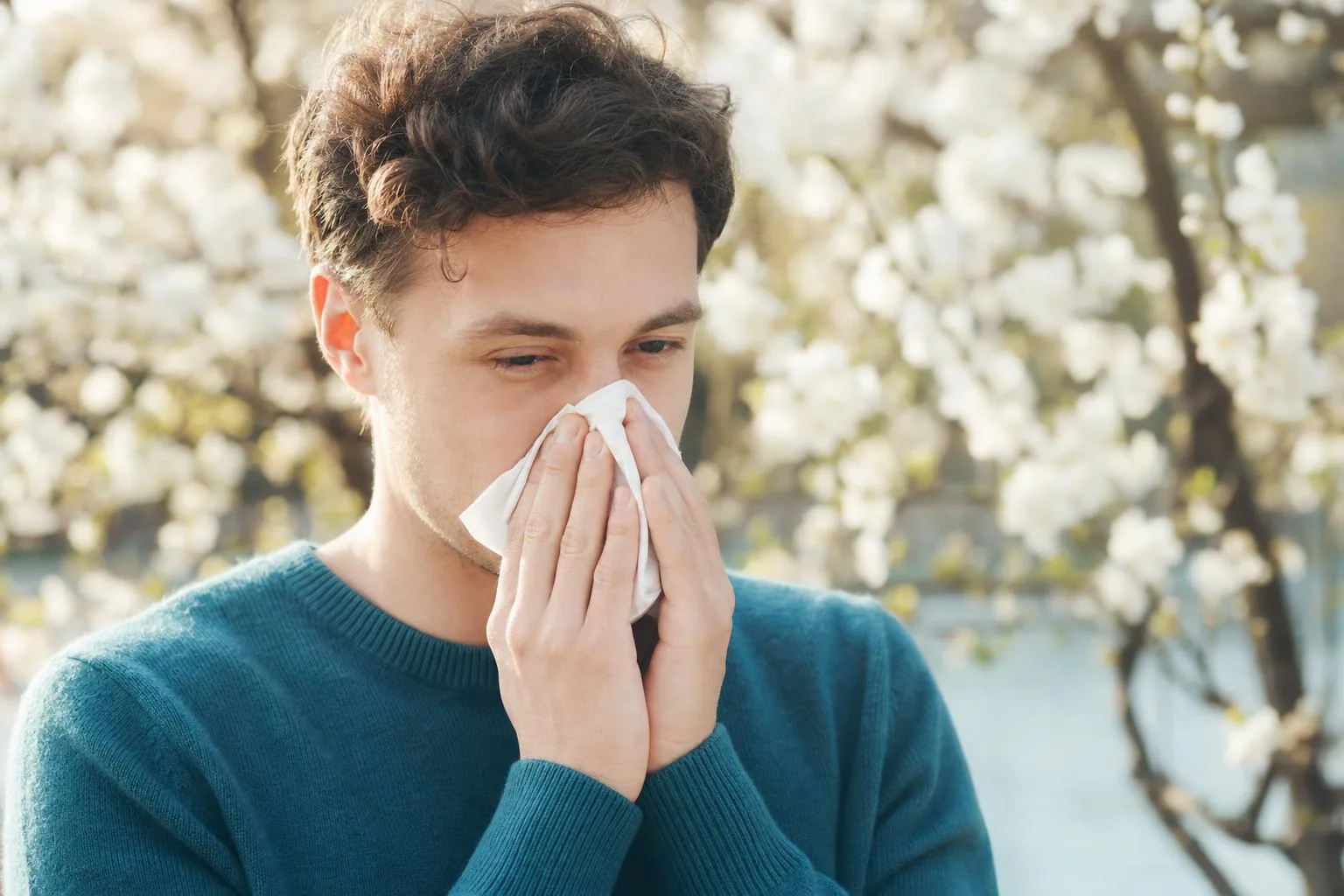 A young adult outdoors in spring pressing a tissue to their nose among blossom